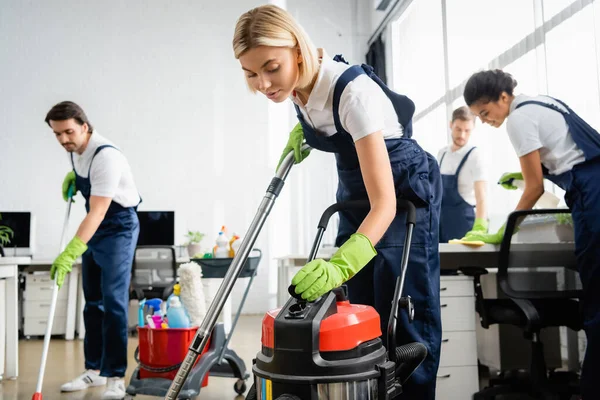 Limpiador en overoles usando aspiradora cerca de colegas multiétnicos en la oficina - foto de stock