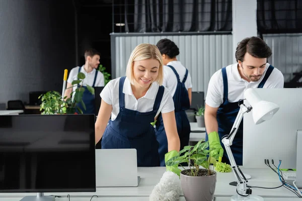 Limpador positivo segurando escova de poeira perto de computadores e colega no escritório — Fotografia de Stock