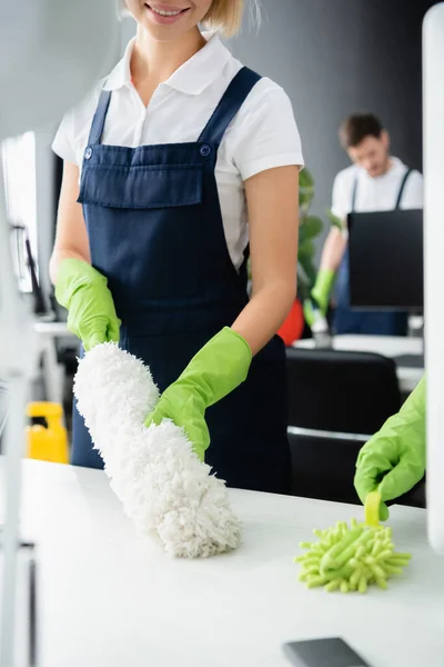 Smiling cleaner in uniform cleaning table with dust brush in office — Stock Photo