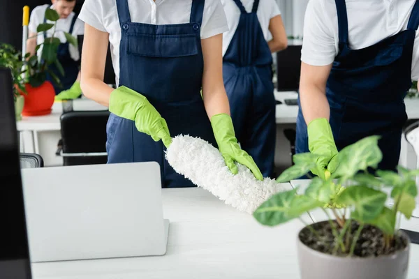 Vista cortada dos trabalhadores do serviço de limpeza segurando escovas de poeira perto da mesa no escritório — Fotografia de Stock