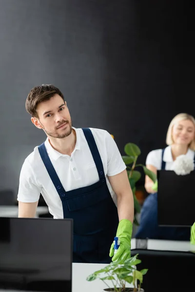 Limpiador con detergente mirando a la cámara cerca de computadoras en la oficina - foto de stock