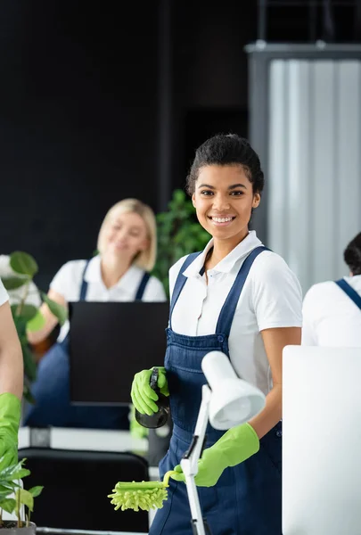 Sonriente limpiador afroamericano con cepillo de polvo y detergente mirando a la cámara en la oficina - foto de stock