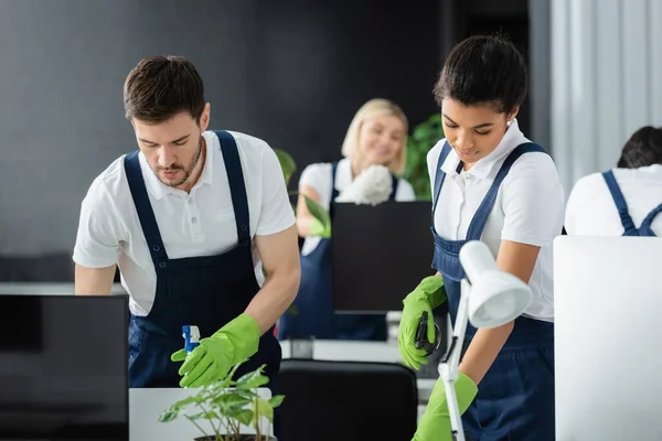 Interracial cleaners with detergents working near computers in office — Stock Photo