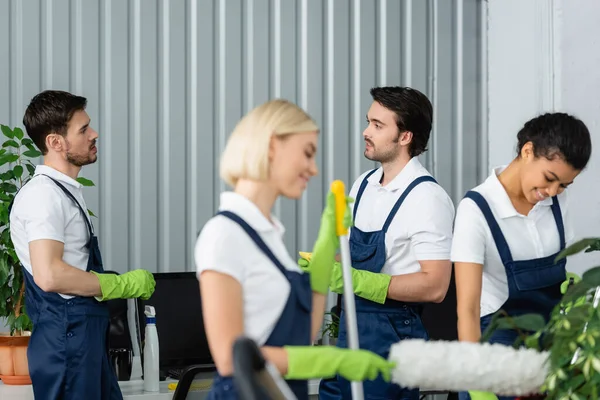 Workers of cleaning service talking near smiling multiethnic colleagues on blurred foreground in office — Stock Photo