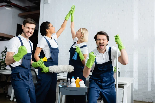 Excited multiethnic cleaners showing yes gesture and high five in office — Stock Photo