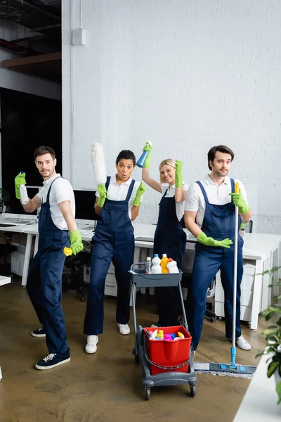 Interracial cleaners in overalls holding cleaning supplies in office — Stock Photo