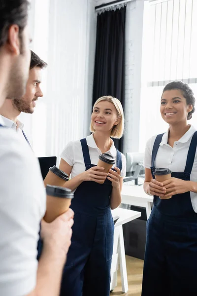 Smiling interracial cleaners with coffee to go looking at colleagues on blurred foreground in office — Stock Photo