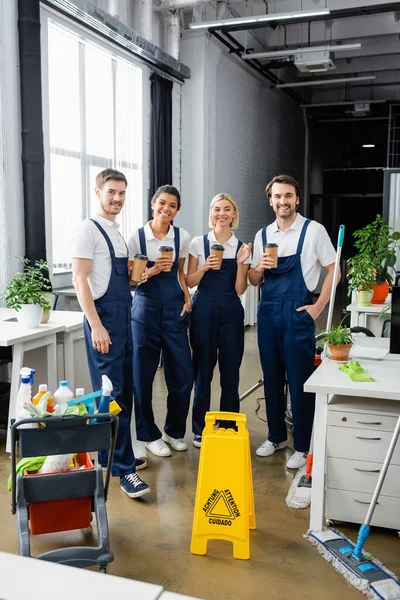 Cheerful multiethnic cleaners holding coffee to go near detergents in office — Stock Photo