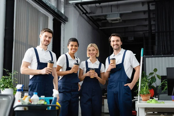 Interracial cleaners with paper cups looking at camera near detergents on blurred foreground in office — Stock Photo