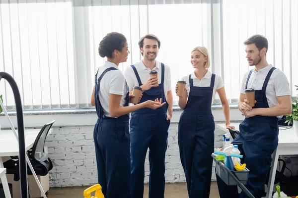 Multiethnic cleaners talking and holding coffee to go in office — Stock Photo