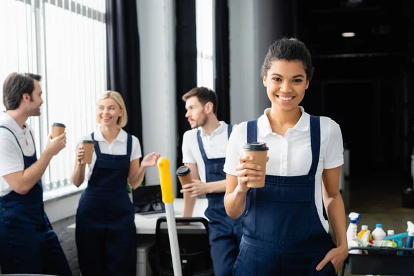 African american cleaner holding paper cup near colleagues on blurred background in office — Stock Photo