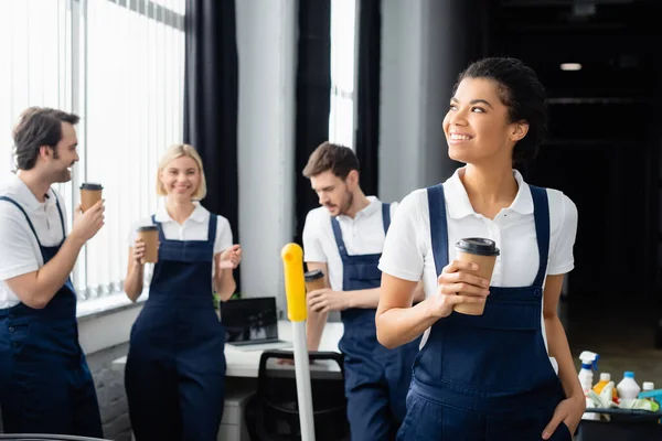 African american cleaner with takeaway drink standing near colleagues talking in office — Stock Photo