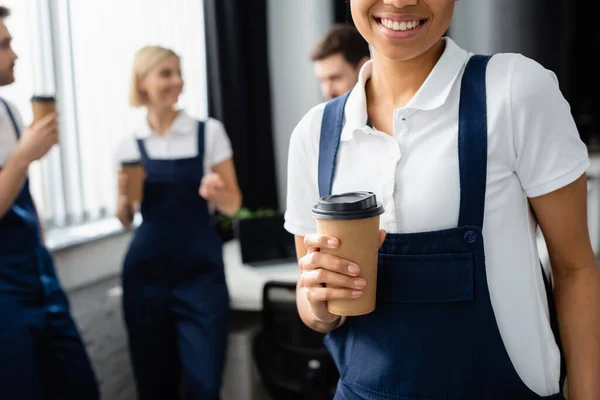 African american cleaner holding paper cup near colleagues on blurred background in office — Stock Photo