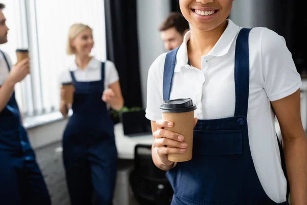 Afrikanische Putzfrau hält Pappbecher neben Kollegen auf verschwommenem Hintergrund im Büro — Stockfoto