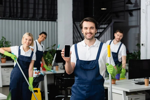 Smiling cleaner holding mop and cellphone with blank screen near multiethnic colleagues in office — Stock Photo