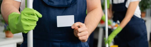 Vue recadrée du nettoyeur avec balai tenant la carte vide dans le bureau, bannière — Photo de stock