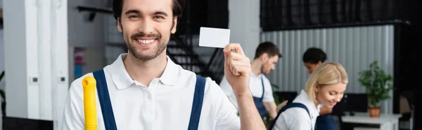 Limpiador sonriente que sostiene la tarjeta vacía cerca de colegas en la oficina, pancarta - foto de stock