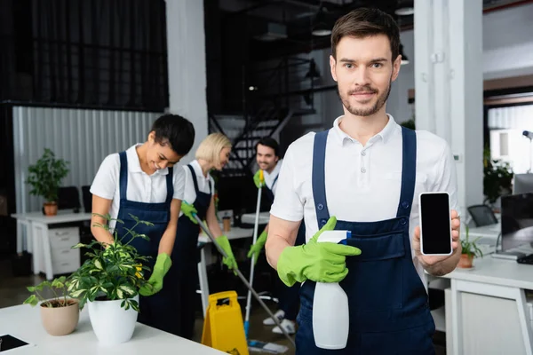 Cleaner with detergent and smartphone standing near multiethnic colleagues on blurred background — Stock Photo