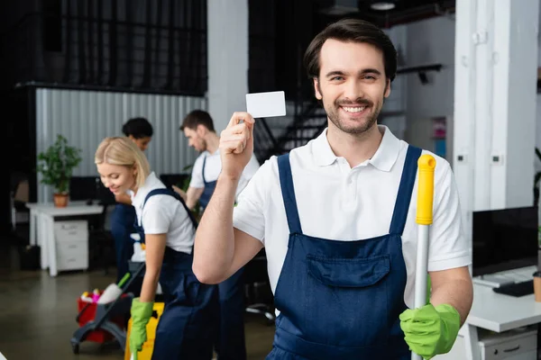 Positive cleaner holding empty card and mop while colleagues working in office — Stock Photo