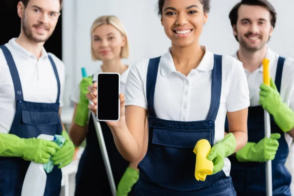 Smartphone en la mano del limpiador afroamericano sonriendo cerca de colegas sobre fondo borroso - foto de stock