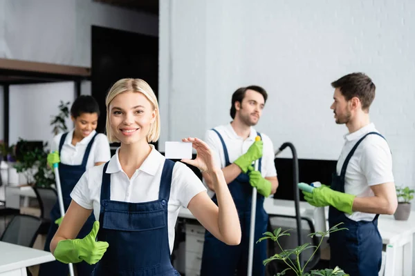 Positive cleaner with empty card showing like near multiethnic colleagues in office — Stock Photo