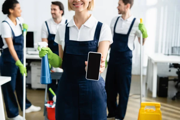 Operaio sorridente di impresa di pulizia che tiene smartphone e detergente su sfondo sfocato in ufficio — Foto stock