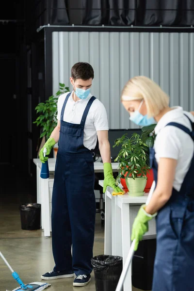 Cleaner in medical mask and uniform cleaning table near colleague on blurred foreground in office — Stock Photo