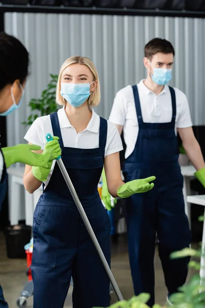 Interracial cleaners in medical masks and uniform standing in office — Stock Photo