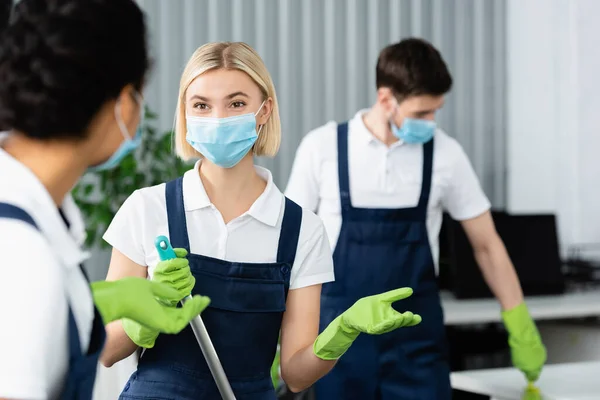 Cleaner em máscara médica segurando esfregona perto de colega afro-americano em primeiro plano desfocado no escritório — Fotografia de Stock