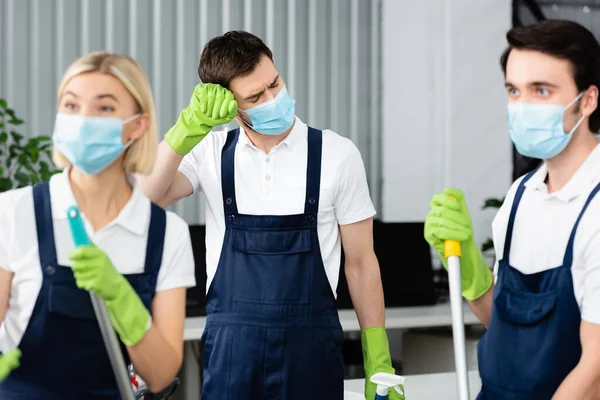 Trabalhador cansado do serviço de limpeza em máscara médica em pé perto de colegas em primeiro plano desfocado no escritório — Fotografia de Stock