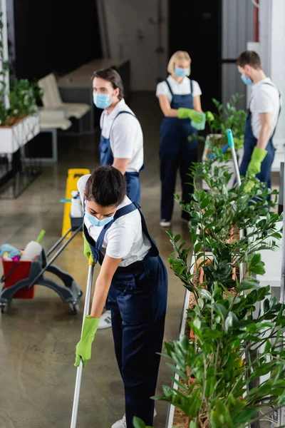 Nettoyant afro-américain au masque médical tenant la serpillière près des plantes au bureau — Photo de stock