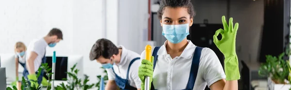 African american cleaner in medical mask showing ok gesture and holding mop in office, banner — Stock Photo