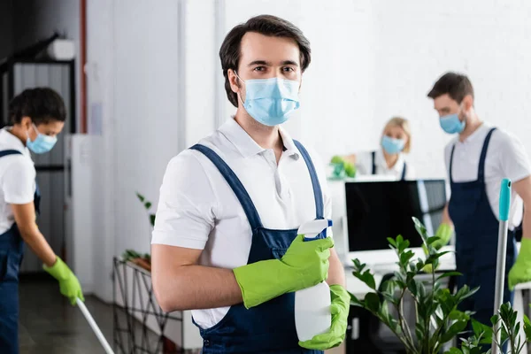 Trabalhador da empresa de limpeza em máscara médica segurando detergente no escritório — Fotografia de Stock
