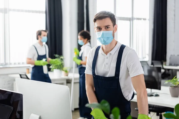 Pulitore in tuta e maschera medica in piedi vicino al computer in ufficio — Foto stock