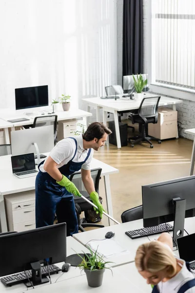 Nettoyant souriant plancher de lavage près des ordinateurs dans le bureau — Photo de stock