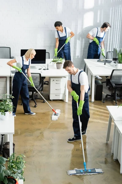 Interracial cleaners in overalls and rubber gloves washing floor in office — Stock Photo