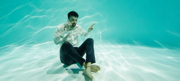 Мусульманский бизнесмен показывает пальцем и разговаривает по смартфону в бассейне, баннер — стоковое фото