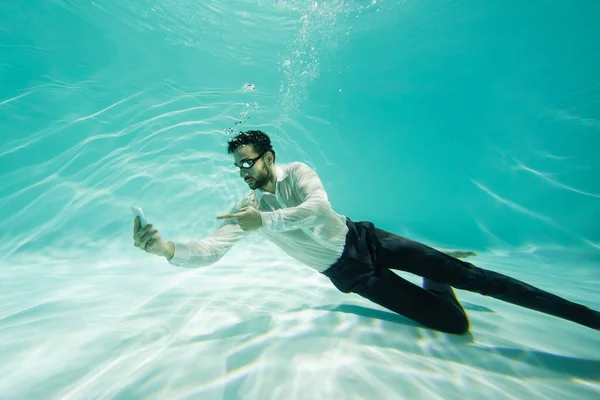 Arabian businessman in goggles pointing at smartphone in swimming pool — Stock Photo