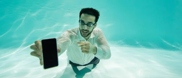 Веселый мусульманский бизнесмен в очках указывает на смартфон с пустым экраном на размытом переднем плане в бассейне, баннер — стоковое фото