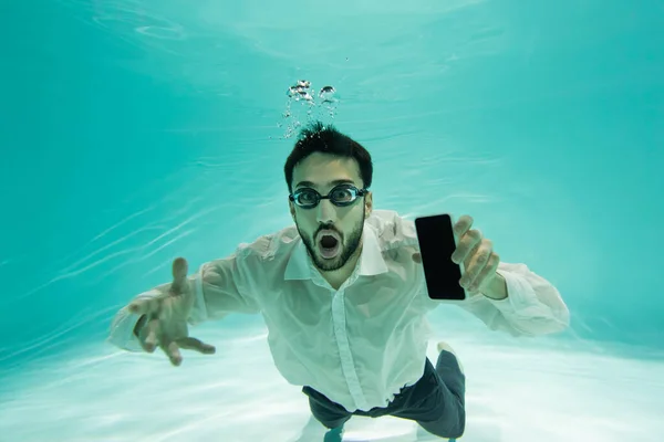 Excited arabian businessman in goggles holding smartphone in swimming pool — Stock Photo