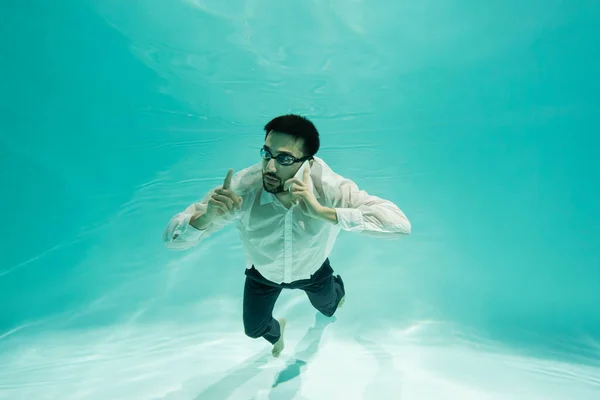 Hombre de negocios musulmán señalando con el dedo mientras habla en el teléfono inteligente bajo el agua - foto de stock