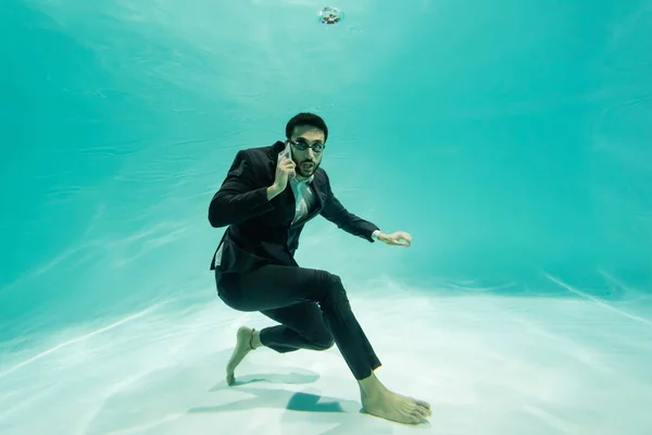Арабский бизнесмен смотрит в камеру, разговаривая на смартфоне под водой в бассейне — стоковое фото