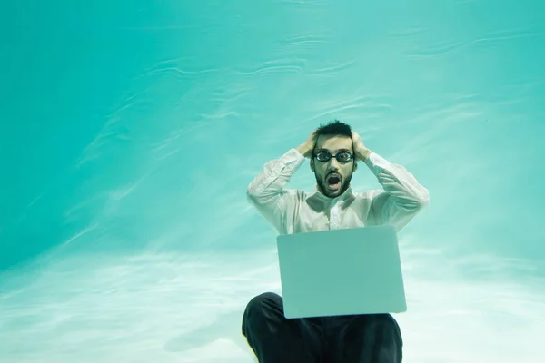 Amazed arabian businessman in goggles looking at camera near laptop in swimming pool — Stock Photo