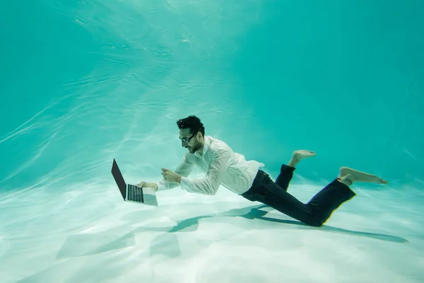 Hombre de negocios árabe sonriente sosteniendo portátil y nadando en la piscina - foto de stock