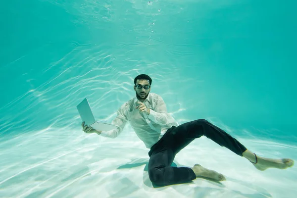 Hombre de negocios musulmán señalando con el dedo mientras sostiene el ordenador portátil en la piscina - foto de stock