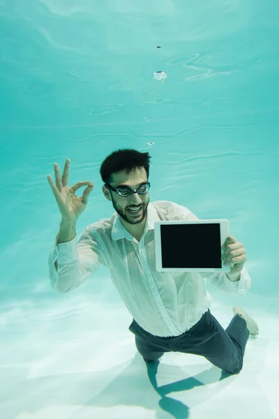 Улыбающийся мусульманский бизнесмен, показывающий нормальный жест и держащий цифровой планшет под водой — стоковое фото