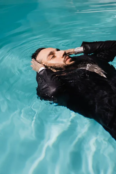 Hombre de negocios árabe con los ojos cerrados nadando en la piscina con agua ondulada - foto de stock