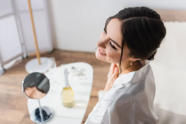 Vista de alto ângulo da noiva feliz sorrindo perto da mesa de cabeceira com espelho e champanhe no fundo borrado — Fotografia de Stock