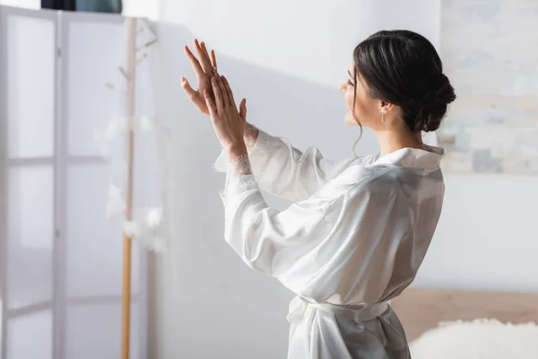 Novia morena en bata de seda mirando anillo de bodas en el dormitorio - foto de stock