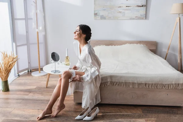 Feliz, novia descalza en bata de seda poniéndose la liga en el dormitorio - foto de stock
